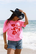 A woman standing on a beach wearing a bright pink t-shirt with a red poppy graphic, denim shorts, and a Roger-Cowboy-Straw-Hat-Black-FleaStyle