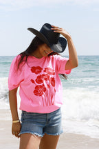 A woman standing on a beach wearing a bright pink t-shirt with a red poppy graphic, denim shorts, and a Roger-Cowboy-Straw-Hat-Black-FleaStyle