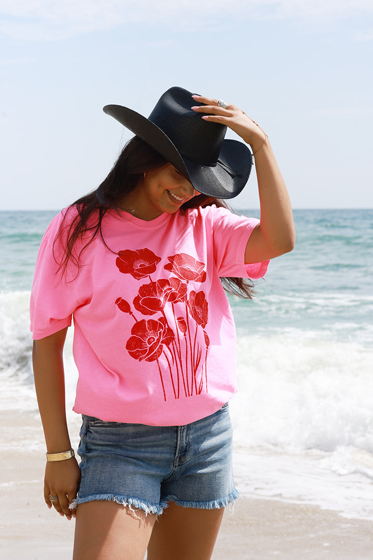 A woman standing on a beach wearing a bright pink t-shirt with a red poppy graphic, denim shorts, and a Roger-Cowboy-Straw-Hat-Black-FleaStyle